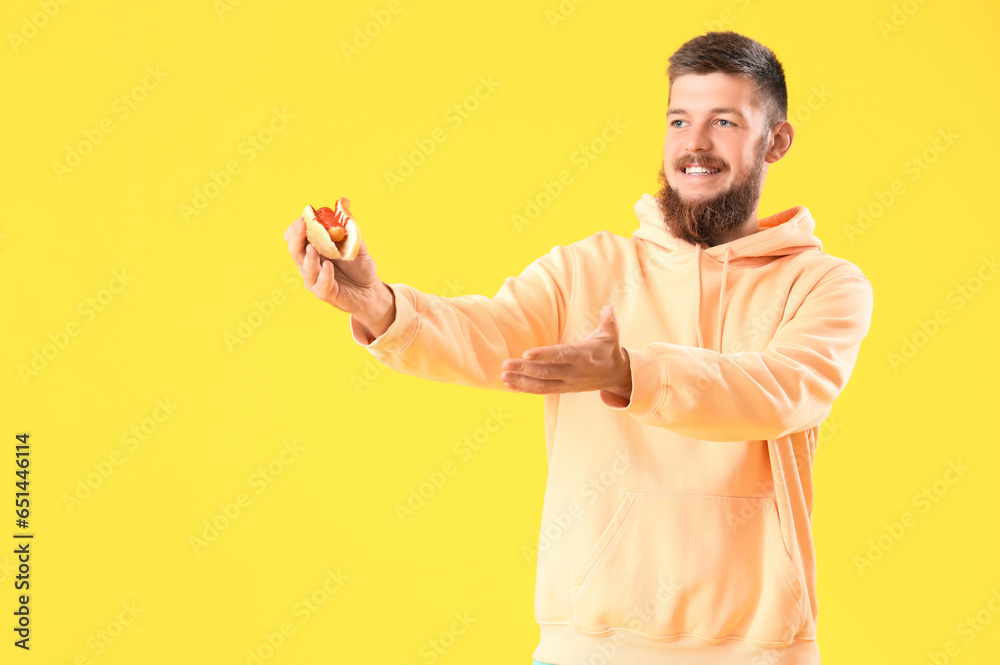 Portrait of happy young man pointing at tasty hot dog on yellow background