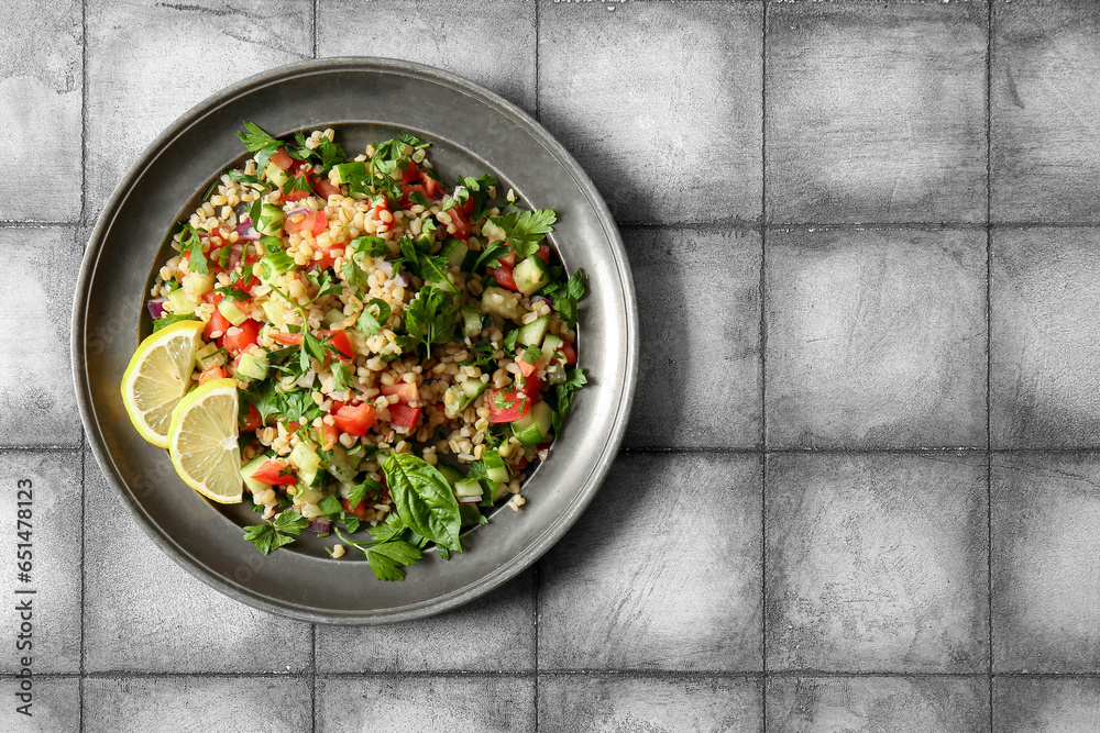 Plate with delicious tabbouleh salad and lemon slices on tile grey background