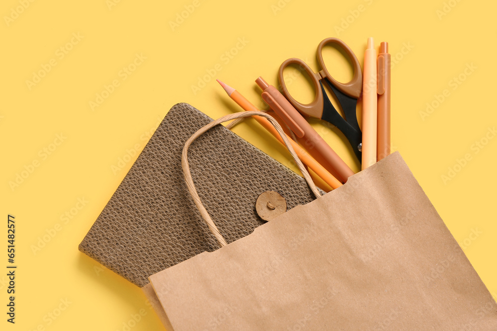 Paper bag with different stationery on yellow background, closeup