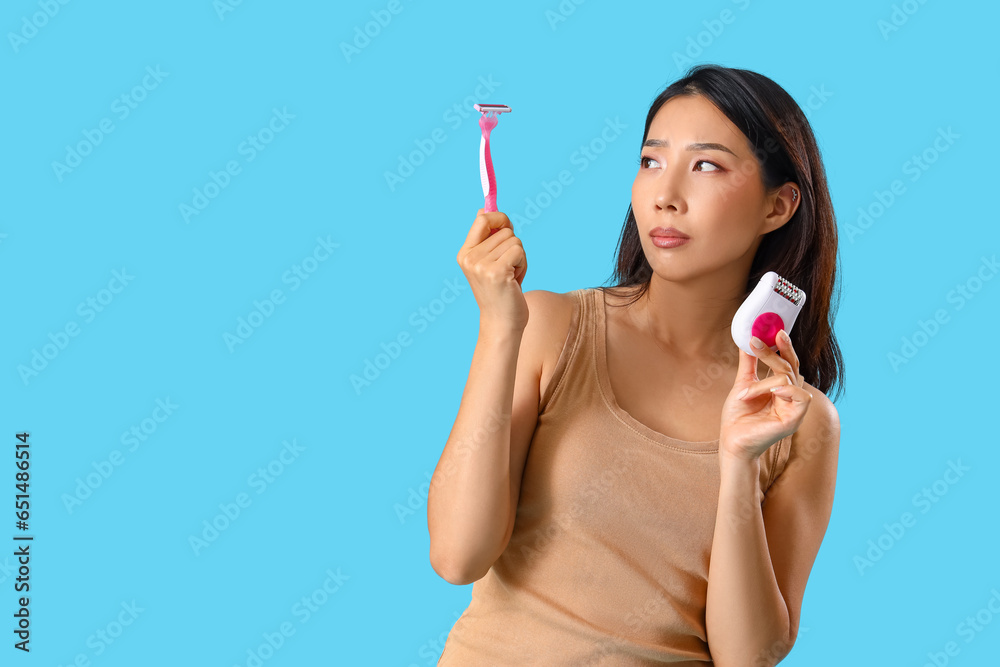 Young Asian woman with razor and epilator on blue background