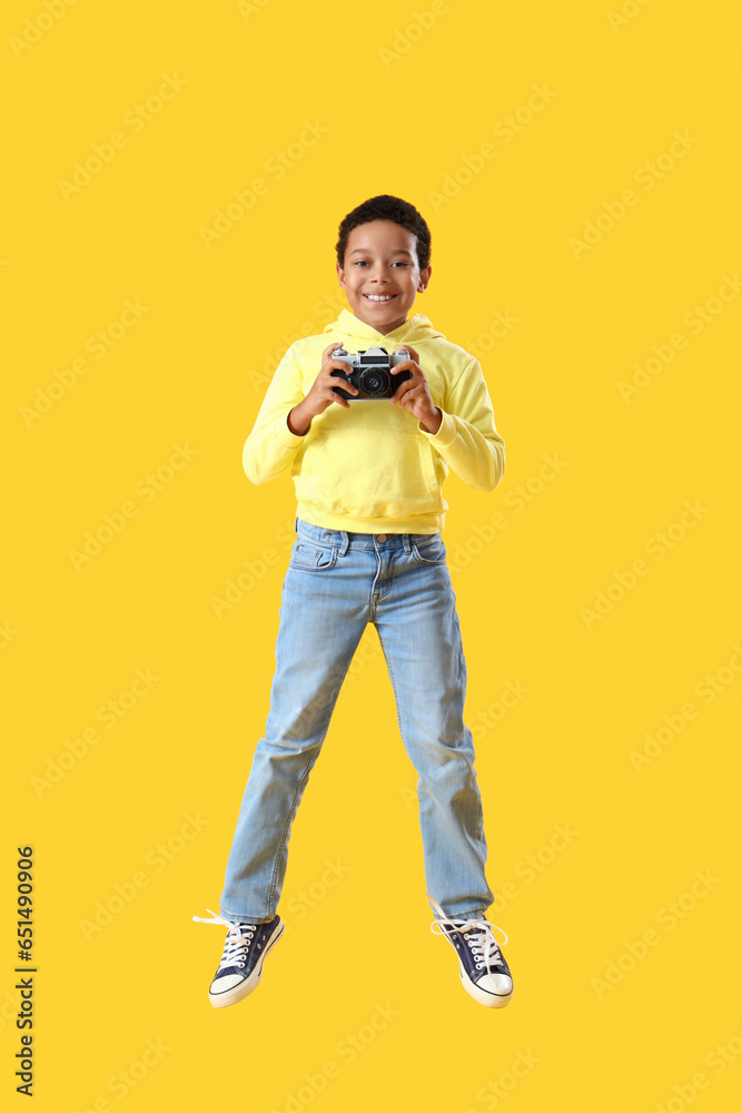 African-American little boy with photo camera jumping on yellow background