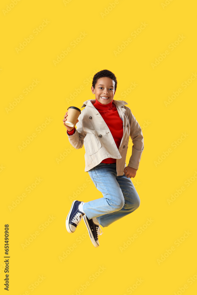 African-American little boy with cup of cocoa jumping on yellow background