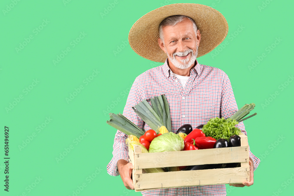 Mature male farmer with wooden box full of different ripe vegetables on green background