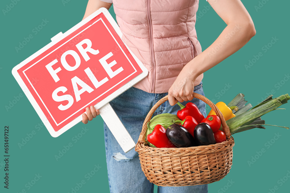 Young female farmer with wicker basket full of different ripe vegetables and FOR SALE sign on green background