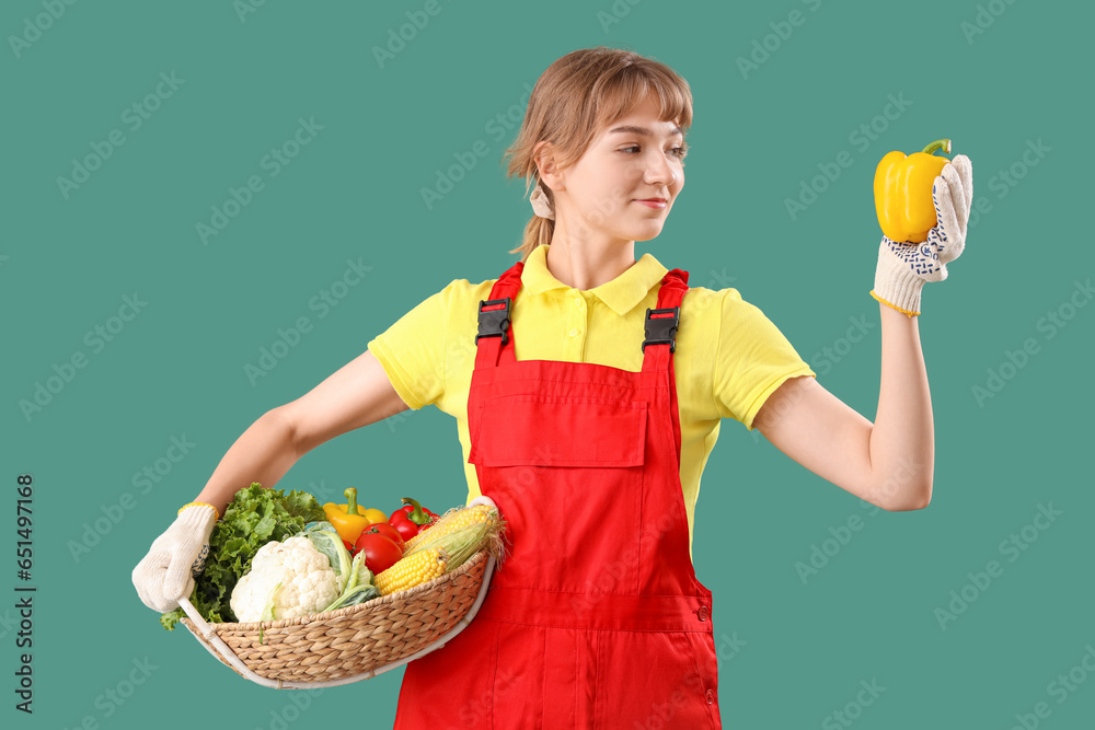 Young female farmer with bell pepper and wicker basket full of different ripe vegetables on green background