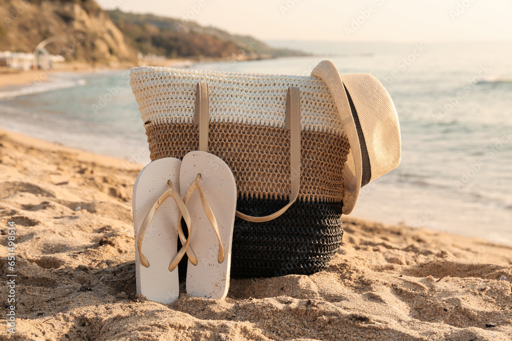 Stylish beach bag, hat and flip-flops on sand near sea