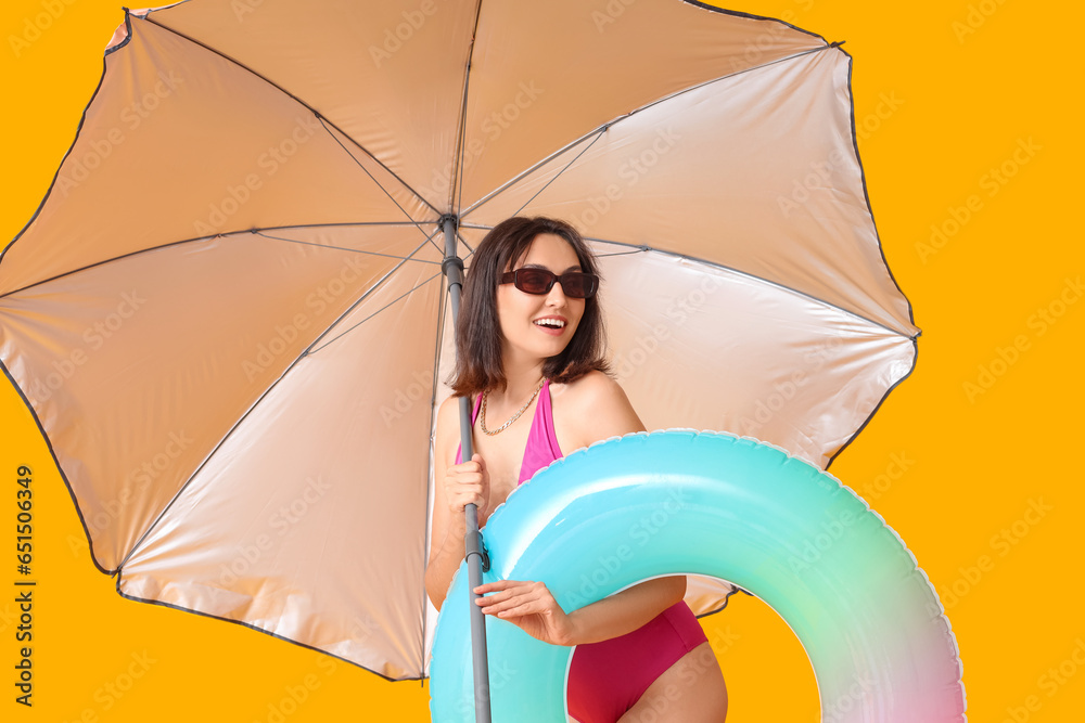 Beautiful young woman with inflatable ring and umbrella on yellow background