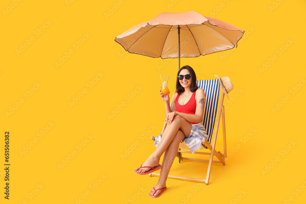 Beautiful young woman with umbrella and glass of tasty cocktail sitting on deckchair against yellow background