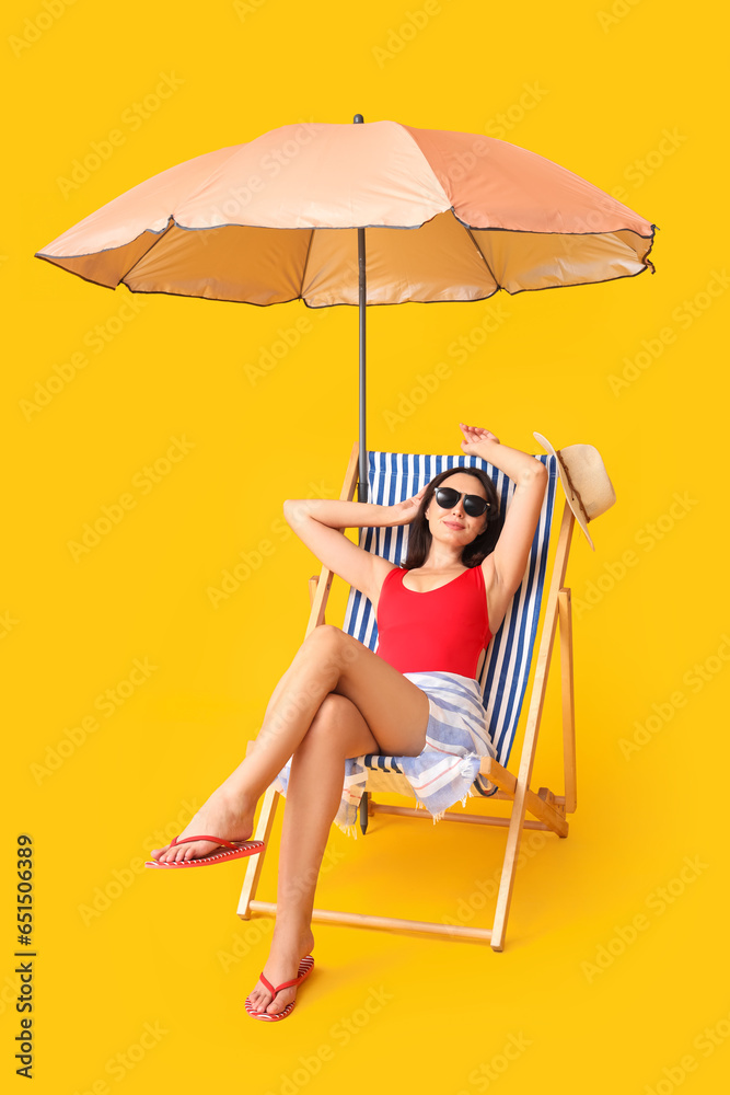 Beautiful young woman with umbrella sitting on deckchair against yellow background