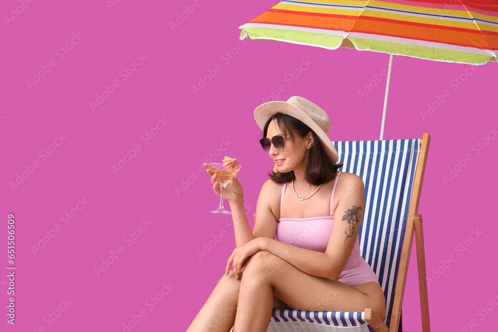 Beautiful young woman with umbrella and glass of tasty cocktail sitting on deckchair against purple background