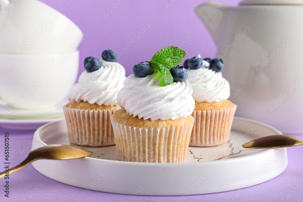 Plate of tasty cupcakes with blueberries on lilac background