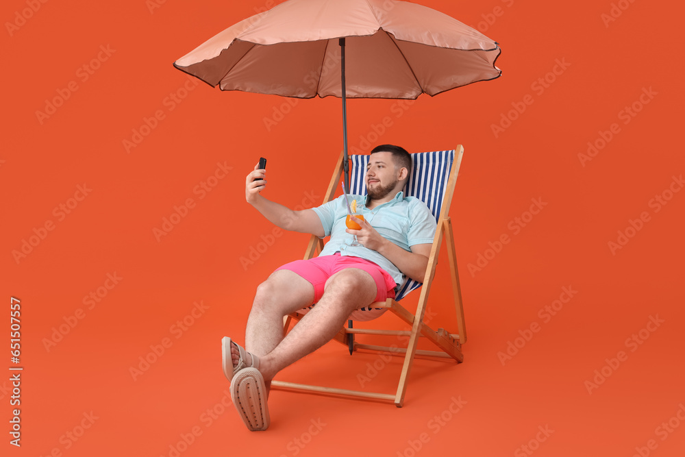 Handsome young man with umbrella, mobile phone and glass of tasty cocktail sitting on deckchair against red background