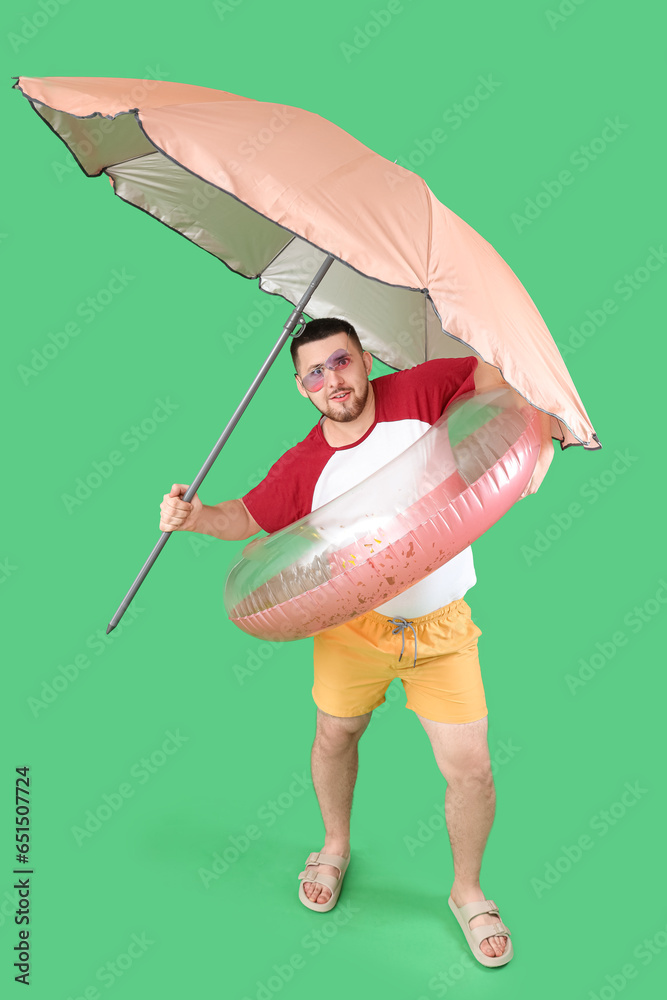 Handsome young man with inflatable ring and umbrella on green background