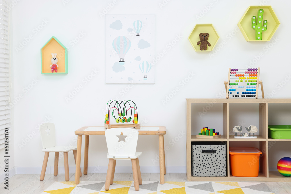 Table with chairs, shelves and toys near white wall in playroom. Stylish kindergarten interior