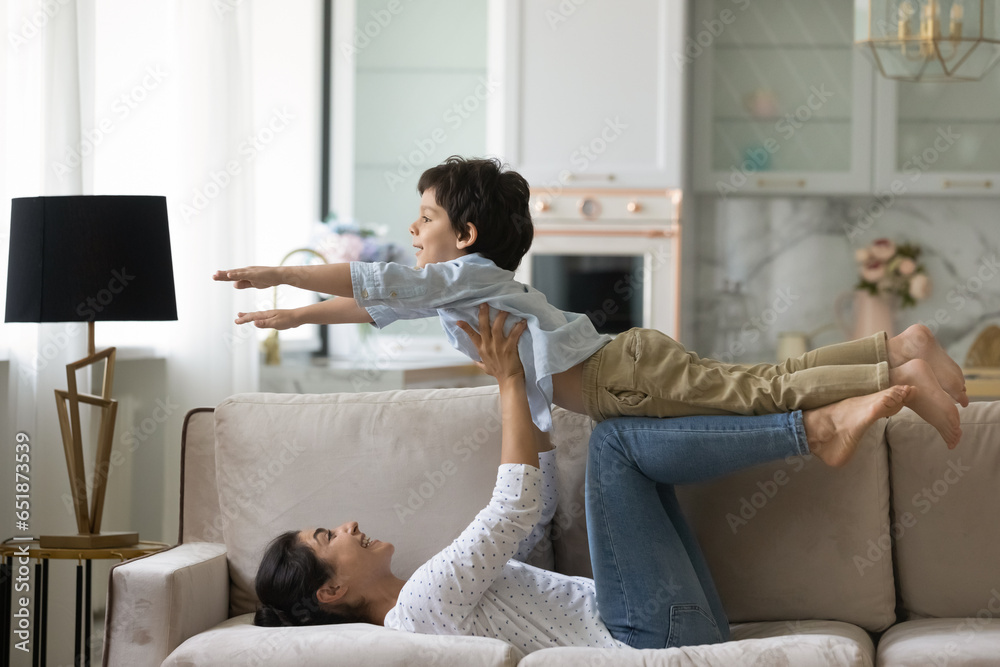 Loving laughing Indian mother lying on couch, lifting 5s son pretending flying super hero, smiling young mom and adorable boy child engaged in funny active game, practicing acroyoga together