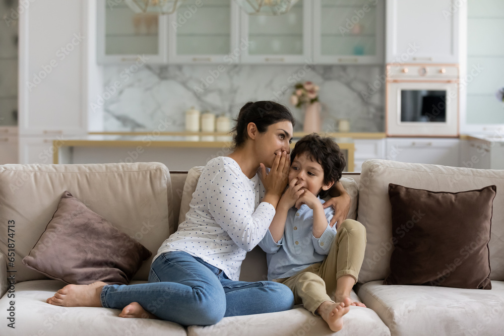 Indian mother whispering in 5s son ear, enjoying pleasant conversation, sharing secrets and gossiping, relaxing sitting on cozy couch at home, spending leisure time, trust and family concept