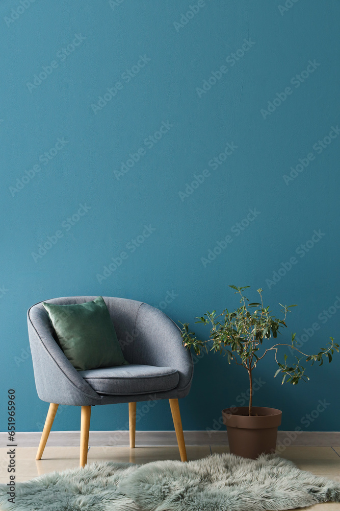 Cozy armchair with cushion, fur rug and houseplant on stool near blue wall