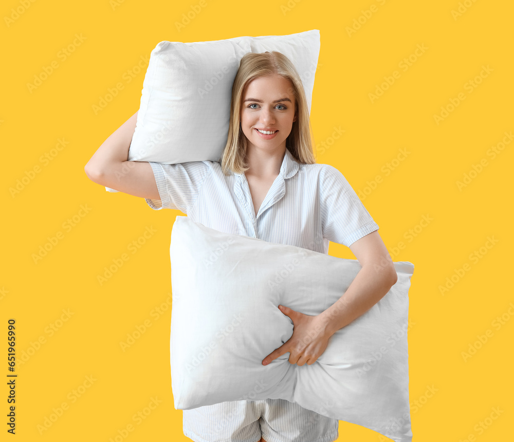 Happy young woman in pajamas and with pillows on yellow background