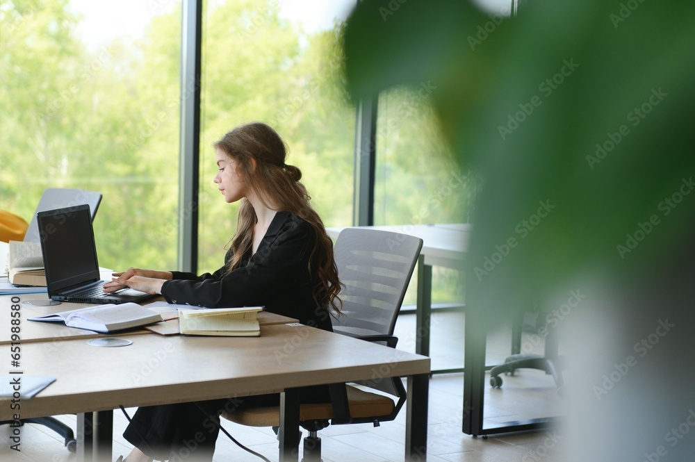 A beautiful caucasian female student is studying in college remotely. She is sitting with a laptop and a notepad and concentrated is watching a video conference lesson