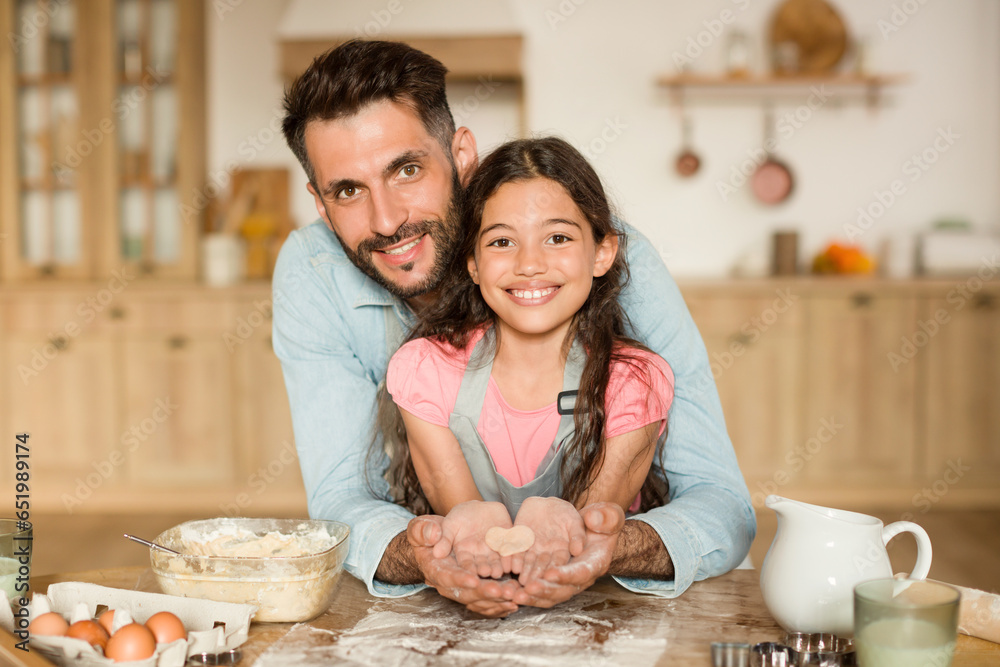 Happy father and daughter holding heart shape made of dough in hands, cooking together in kitchen and smiling at camera