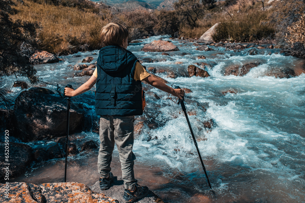 Boy hiker with trekking poles standing on stone at the mountain river in autumn