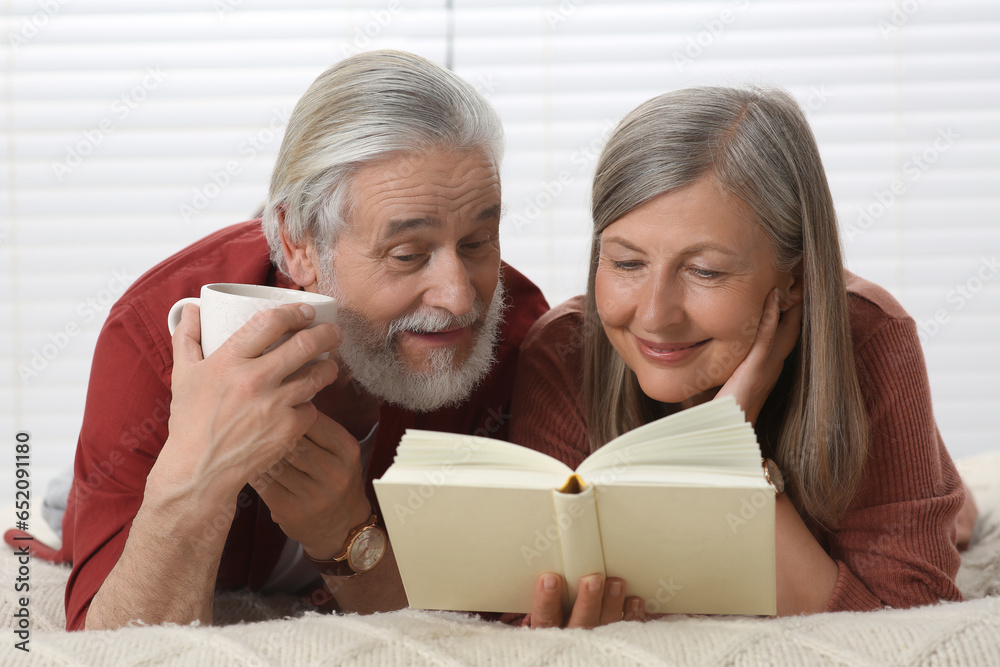 Senior man with cup of drink and his wife reading book on bed at home