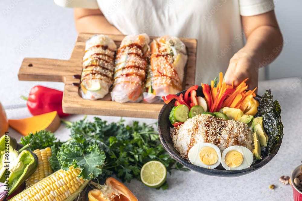 Bowl with vegetables and rice and spring rolls in female hands in the kitchen.