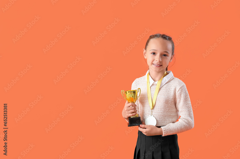 Smart little schoolgirl in stylish uniform with prize cup and medal on orange background