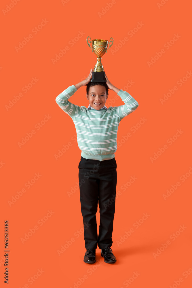 Smart African-American schoolboy in stylish uniform with prize cup on orange background