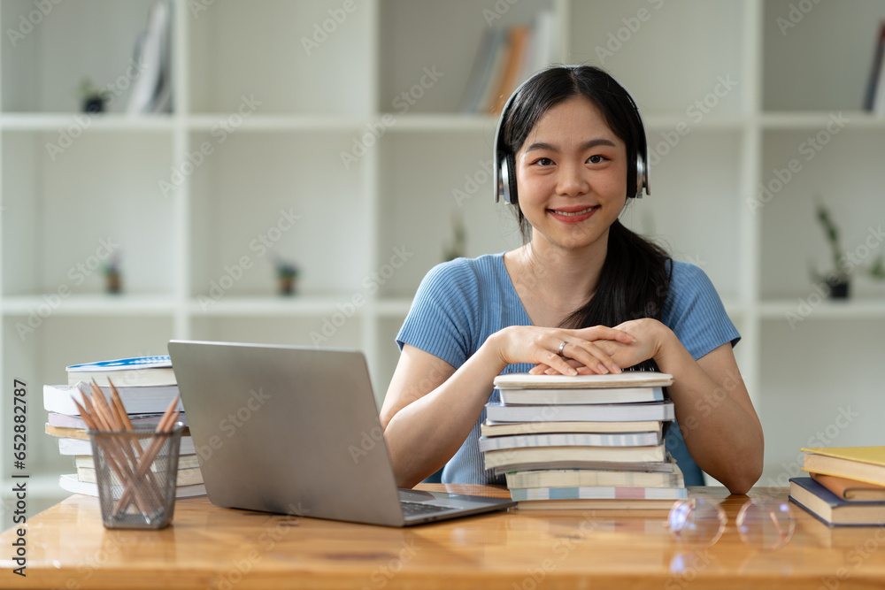 Asian woman in headphones sitting on stack of books and laptop researching and relaxing listening to music while sitting on desk at home on weekends