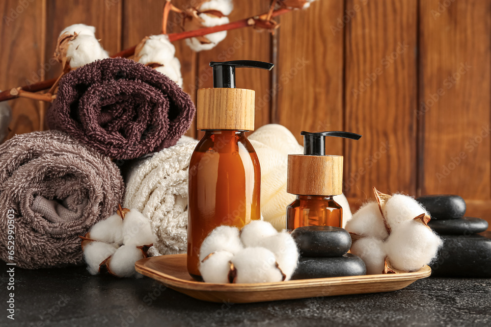 Composition with bottles of cosmetic products, bath supplies and cotton flowers on dark table against wooden background, closeup