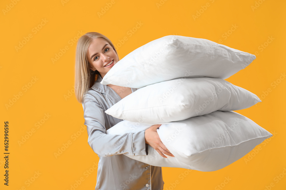 Young woman in pajamas and with pillows on yellow background