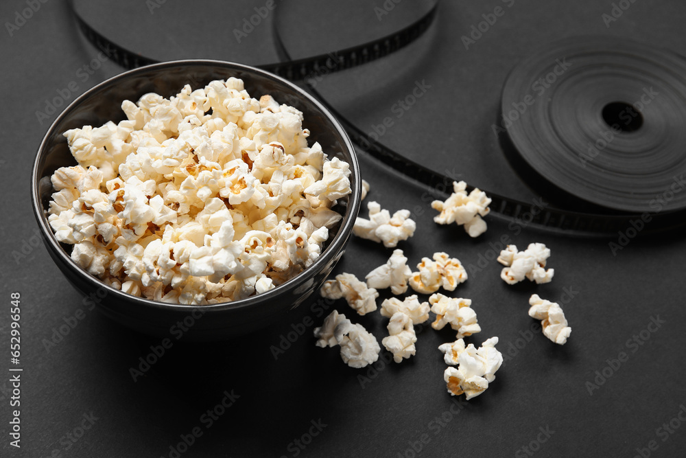 Bowl with tasty popcorn and film reel on black background