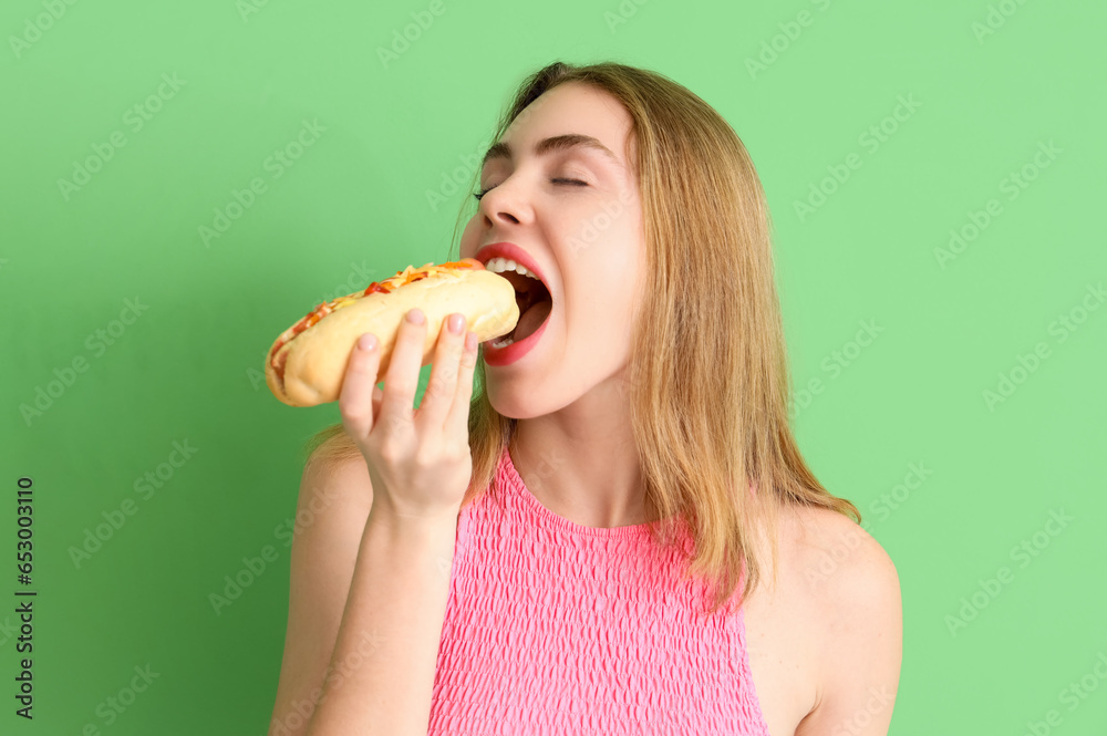 Pretty young woman eating tasty hot dog on green background