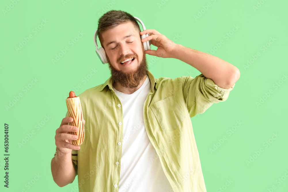 Portrait of happy young man with tasty hot dog listening to music on green background