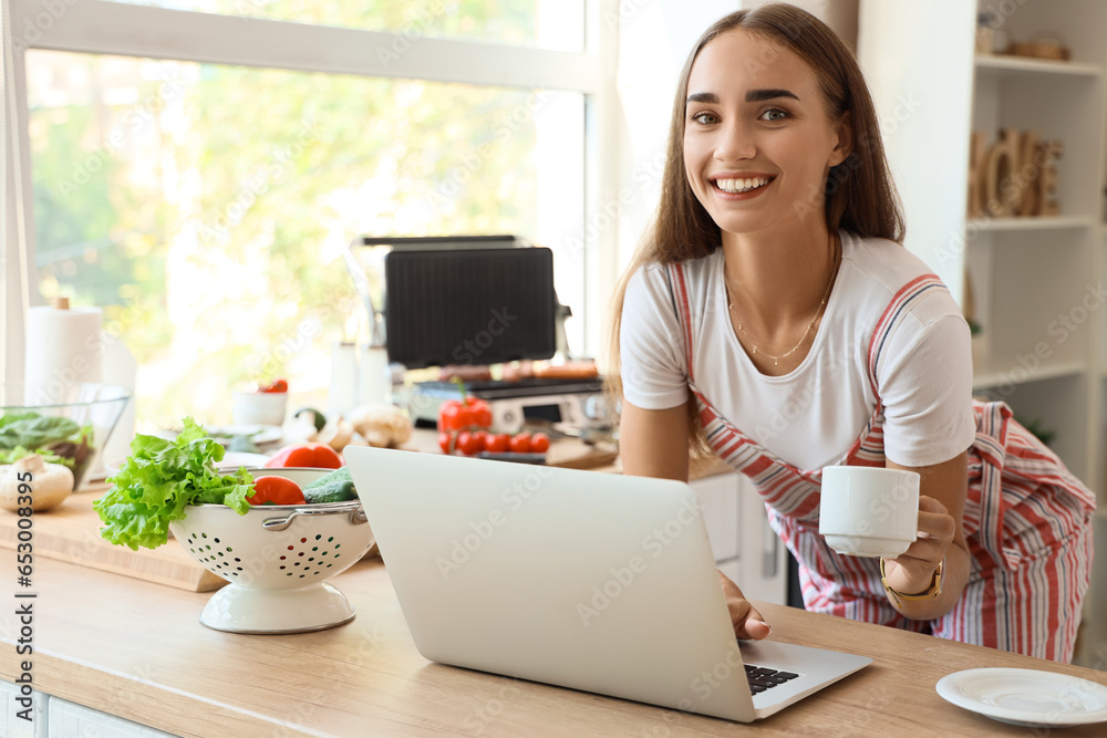 Beautiful young woman using laptop while cooking delicious sausages on modern electric grill in kitchen