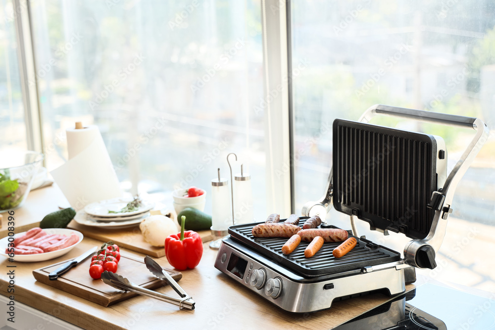 Modern electric grill with tasty sausages on wooden countertop in kitchen