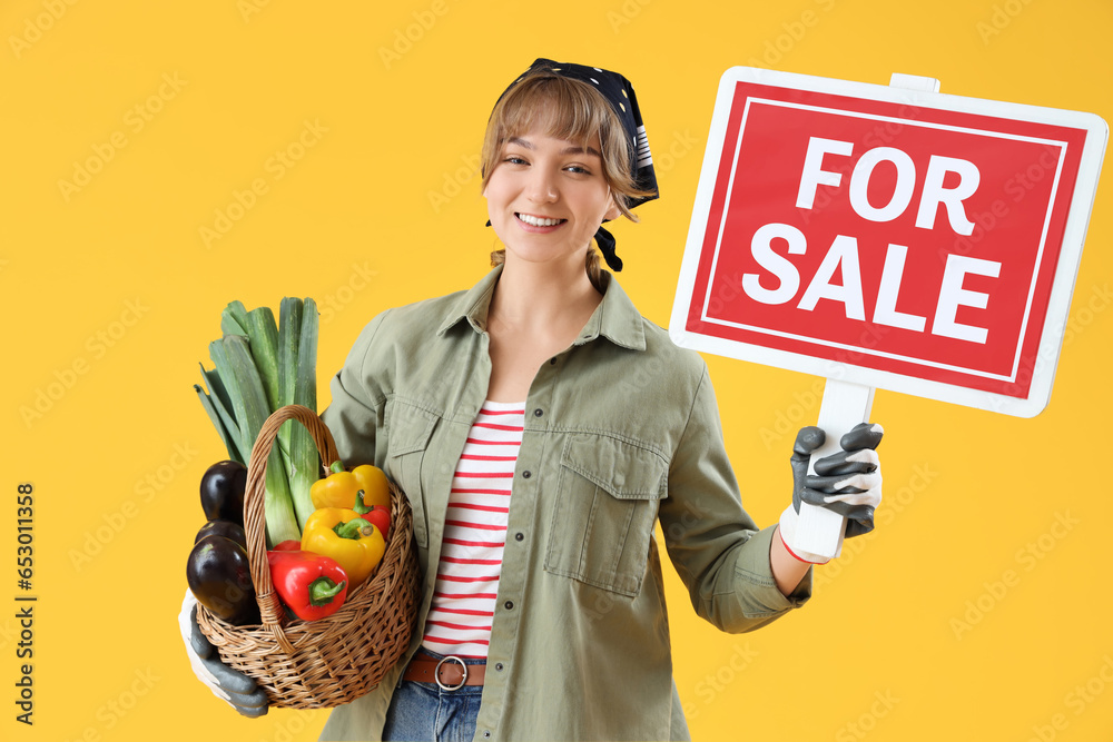 Young female farmer with wicker basket full of different ripe vegetables and FOR SALE sign on yellow background