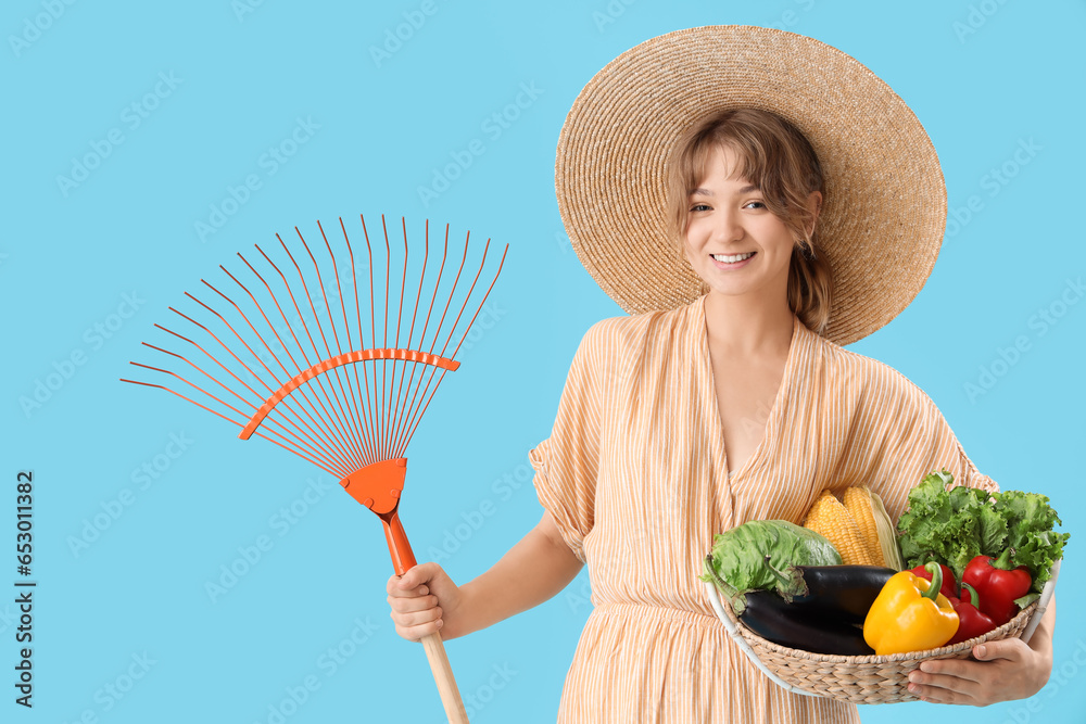 Happy young female farmer with rake and wicker basket full of different ripe vegetables on blue background