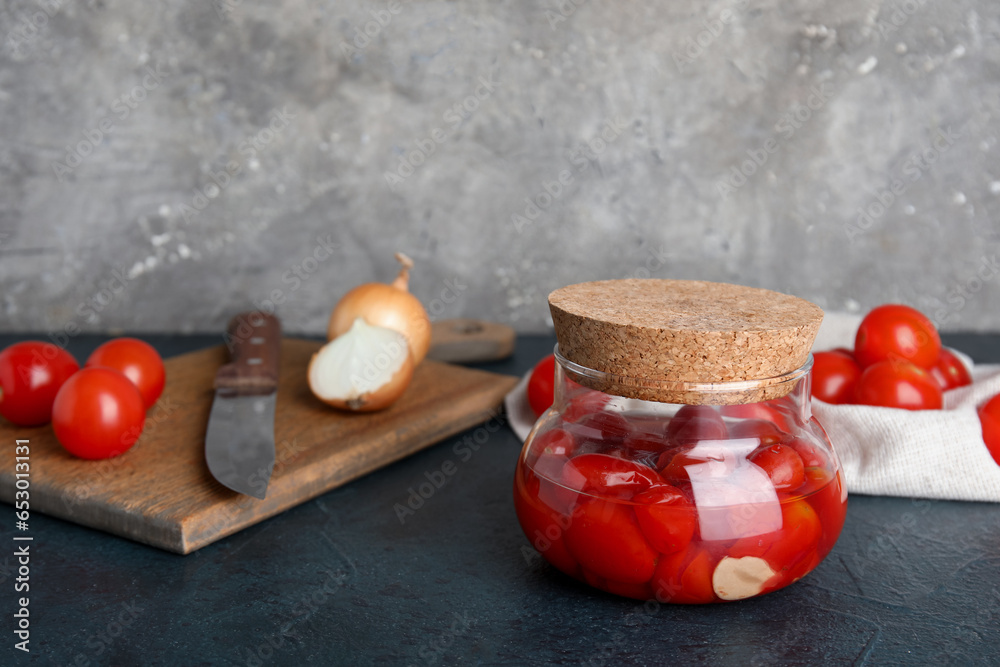 Jar of pickled tomatoes and wooden board with onion on dark table