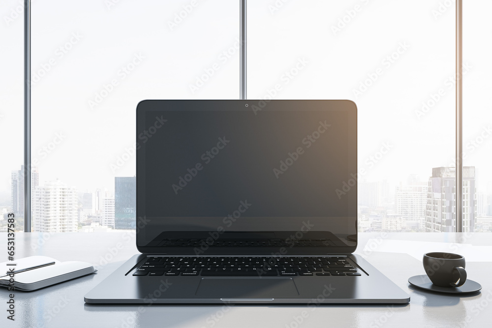 Close up of modern workplace with empty mock up laptop screen, supplies, coffee cup and panoramic window with city view in the background. 3D Rendering.