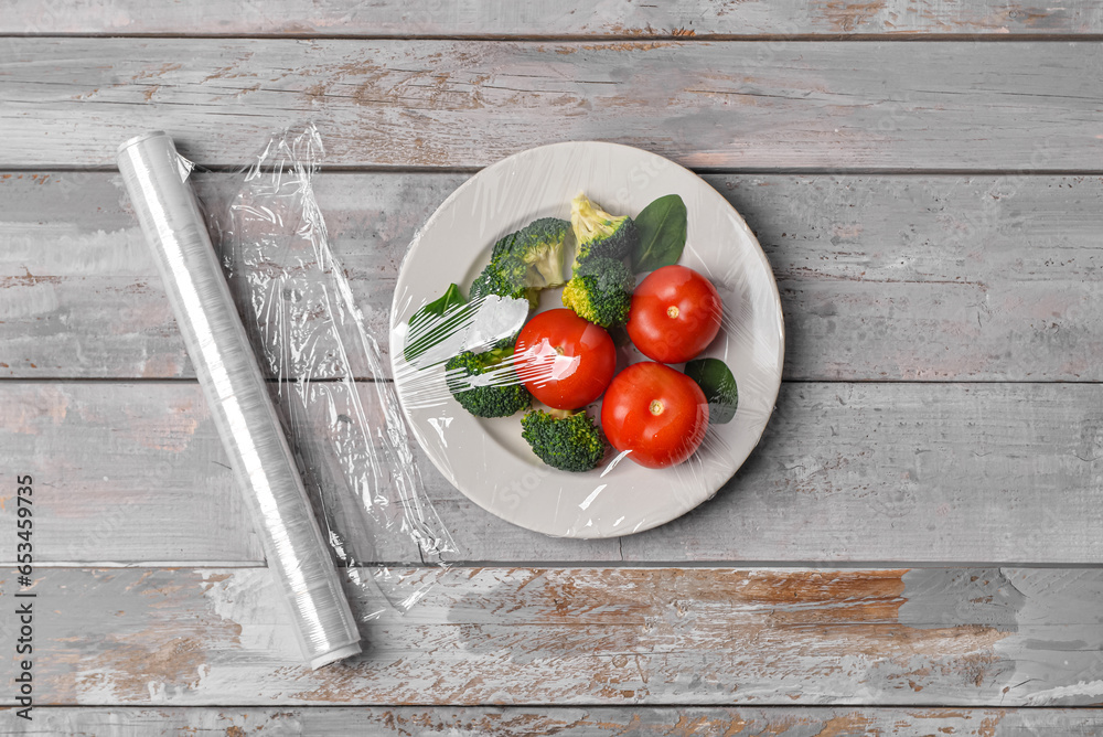 Plate with vegetables wrapped with stretch wrap on grey wooden background