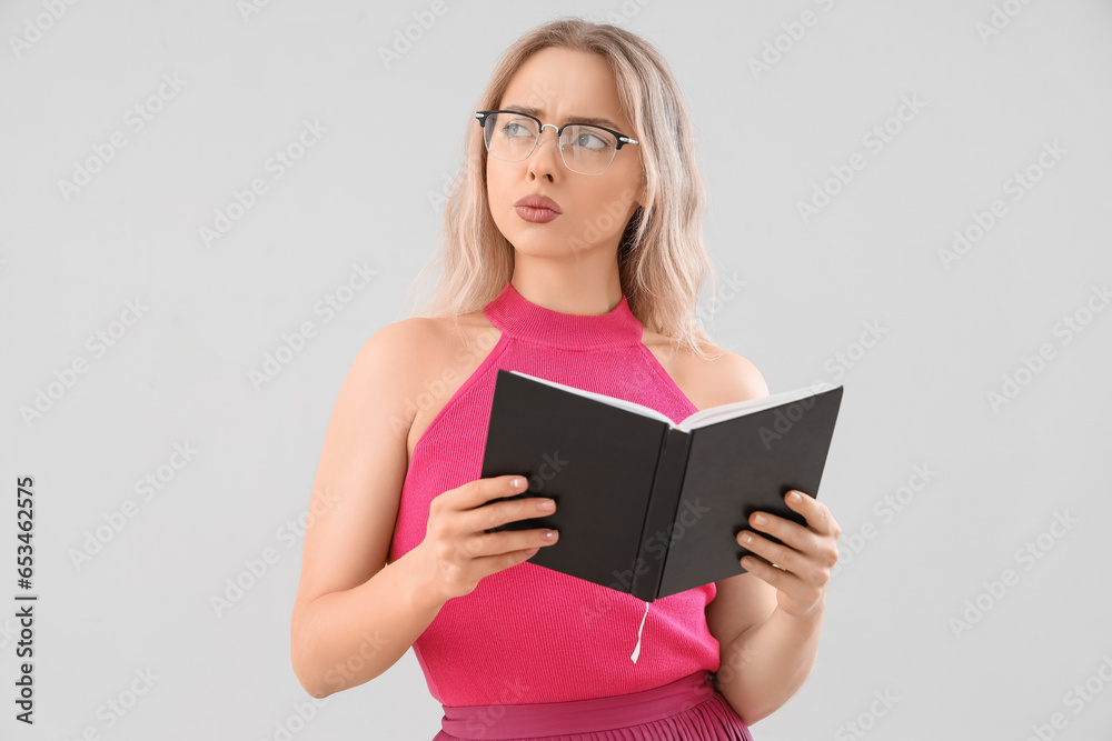 Thoughtful young woman in stylish eyeglasses reading book on light background