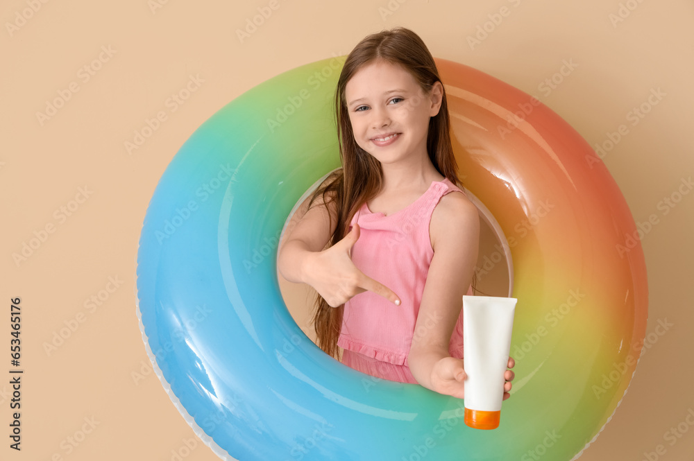 Little girl with inflatable ring pointing at bottle of sunscreen cream on beige background