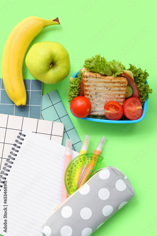 Different stationery and lunch box with tasty food on green background