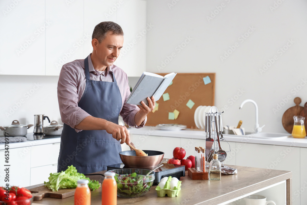 Mature man frying vegetables with recipe book in kitchen