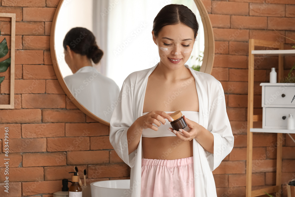 Beautiful Asian woman with jar of cream in bathroom