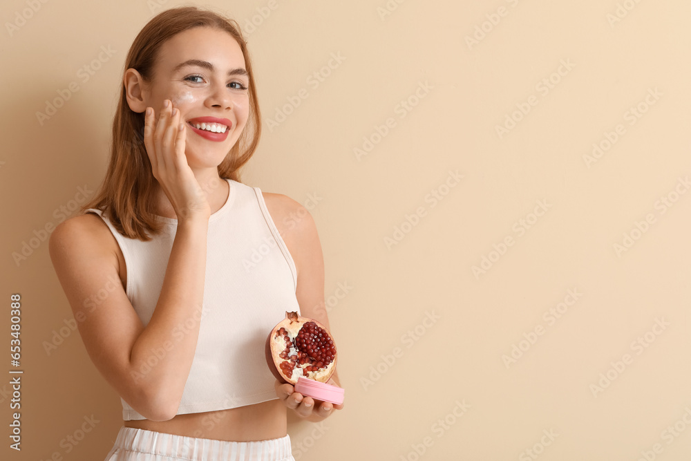 Young woman with pomegranate applying cream on beige background