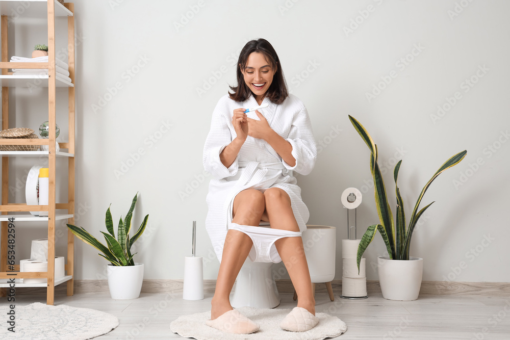 Young woman with pregnancy test sitting on toilet bowl in restroom