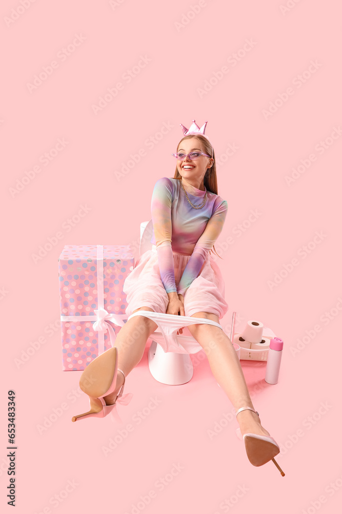 Young woman sitting on toilet bowl and birthday gift against pink background